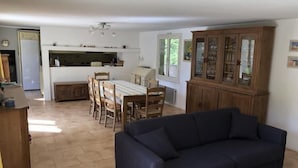 A living room with a dark blue sofa, a dining area with wooden chairs and a table, and a kitchen with white cabinets and a window. - La Téolière (countryside and private pool) (FIGANIERES)