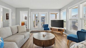 A living room with a grey sofa, blue chairs, a round coffee table, and a TV on a wooden stand. - Tee Off by Brunswick Vacation Rentals: 6 Bedrooms with Amazing Views & Pool! (Ocean Isle Beach)