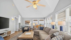 A modern living room with a ceiling fan, a flat-screen TV, a sofa, a coffee table, and a dining area with a table and chairs. - Tee Off by Brunswick Vacation Rentals: 6 Bedrooms with Amazing Views & Pool! (Ocean Isle Beach)