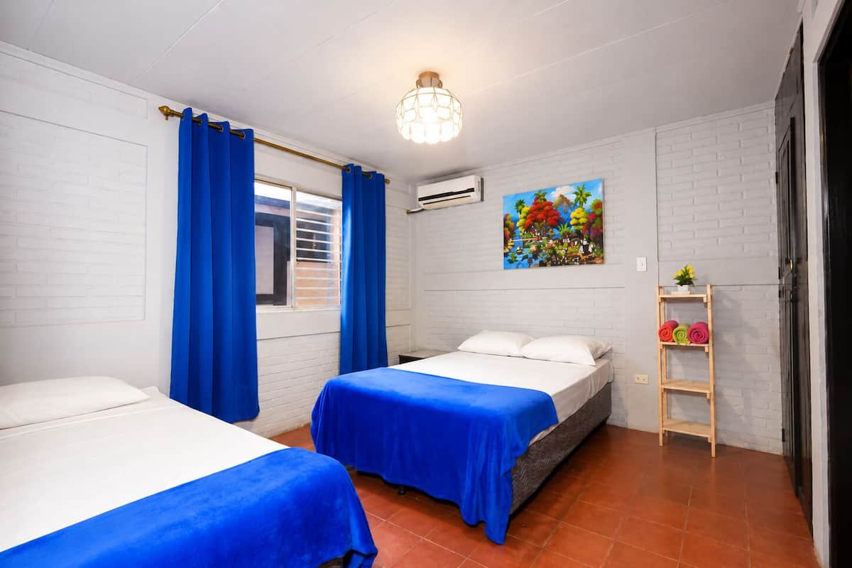 A hotel room with two beds, a window with blue curtains, a wall-mounted air conditioner, a painting, and a wooden shelf with towels.