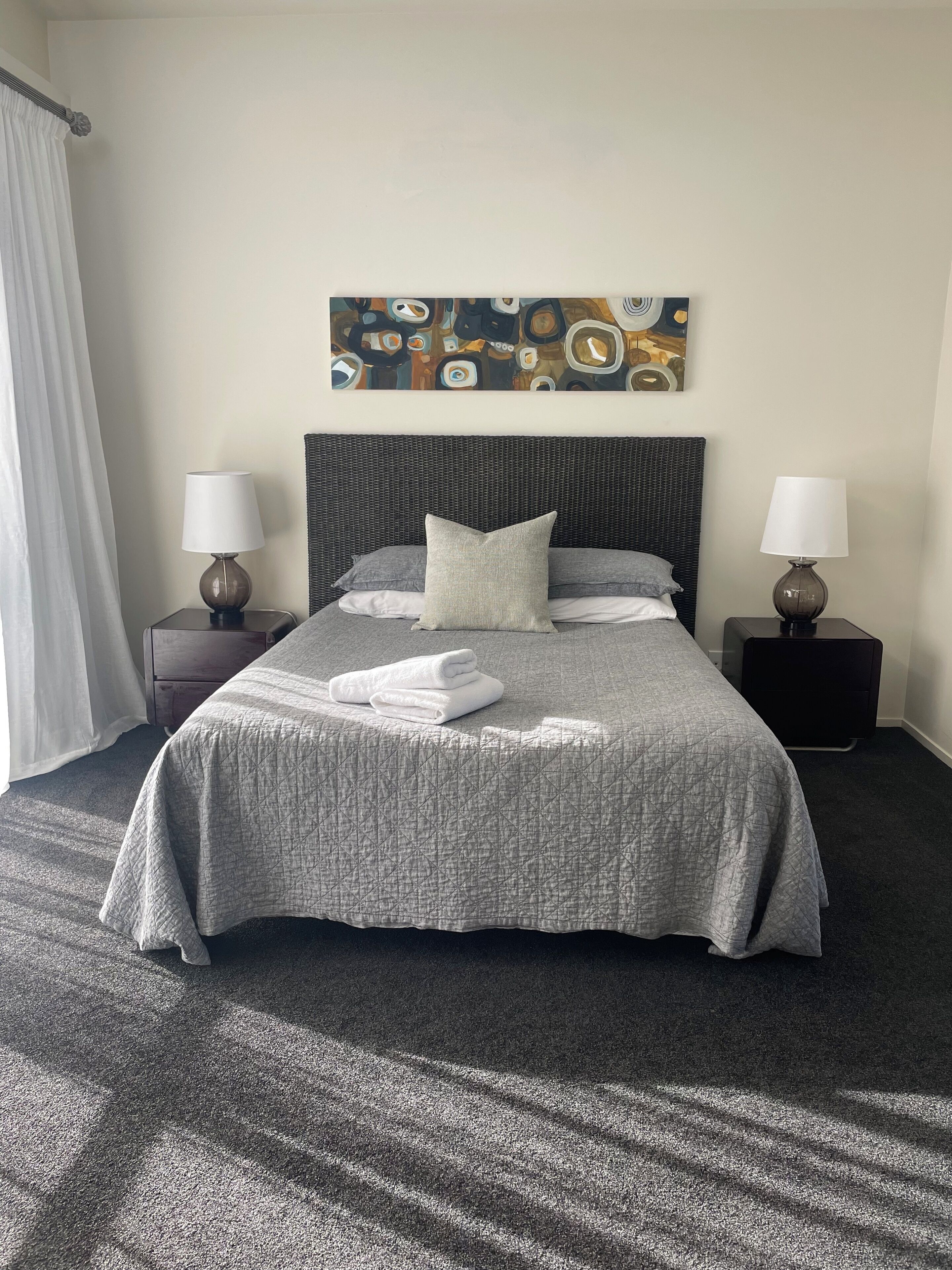 A neatly made bed with a grey quilt, two bedside tables with lamps, a large abstract painting on the wall, and a window with white curtains.