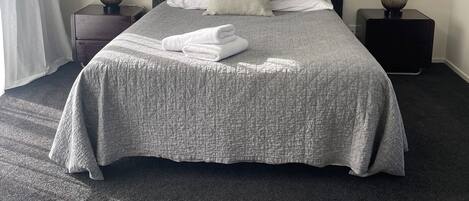 A neatly made bed with a grey quilt, two bedside tables with lamps, a large abstract painting on the wall, and a window with white curtains.