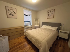 Un dormitorio con cama, cómoda, mesita de noche, ventana con persianas y dos cuadros florales en la pared.