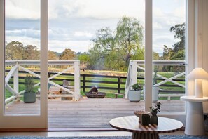 A deck with a view of a grassy field and trees, a wooden chair, a fire pit, and a table with a lamp. - Farm Stay Feel, Tiny Home Vibes (Merton)