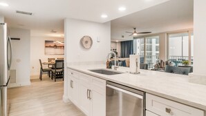 Une cuisine moderne équipée d’un lave-vaisselle, un coin repas avec une table et des chaises, et une vue sur le salon avec un canapé et un balcon.