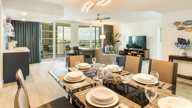 Un espace repas moderne avec une table en verre pouvant accueillir six personnes, un téléviseur et un balcon offrant une vue agréable.