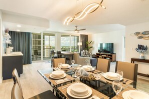 A modern dining area with a glass table set for six, a television, and a balcony with a view. - Tide & Seek | Beach-front and Resort Amenities (Fort Myers Beach)