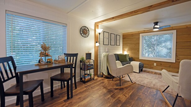 Un salon chaleureux avec un plancher en bois, une table à manger avec des chaises, un fauteuil à bascule, un canapé et une grande fenêtre avec des stores.