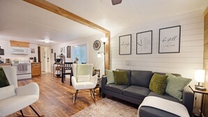 Un salon moderne avec un canapé gris, un fauteuil blanc et un fauteuil à bascule. Le sol est en bois, il y a un petit tapis et, au fond, on aperçoit un coin repas.