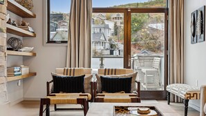 A cozy living room with wooden furniture, a large window with a view of houses, and a bookshelf with decorative items.