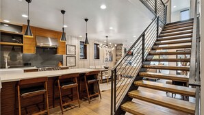 A modern kitchen with a central island, bar stools, pendant lighting, and a staircase with wooden steps and metal railings.