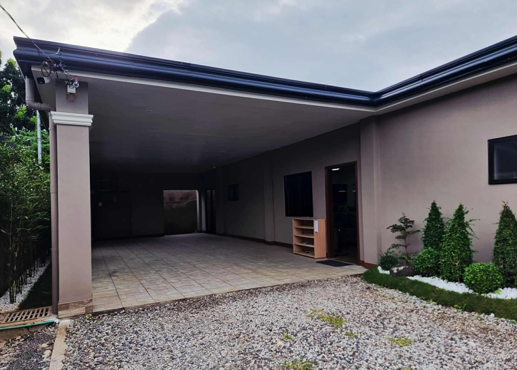 A covered parking area with a gravel driveway leading to a building with a flat roof and a small window.