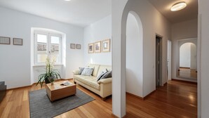 Ein modernes Wohnzimmer mit Holzboden, einem beigefarbenen Sofa, einem Couchtisch und einem Topf mit Pflanze. An der Wand hängen Bilderrahmen und das Fenster bietet einen Blick auf Gebäude.