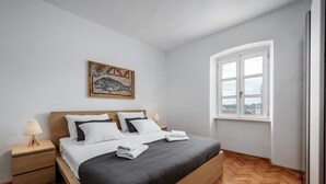 Ein ordentlich bezogenes Bett mit dunkler Decke, einem Holzkopfende und zwei Nachttischen mit Lampen. Rechts ist ein Fenster mit weißen Rahmen zu sehen.