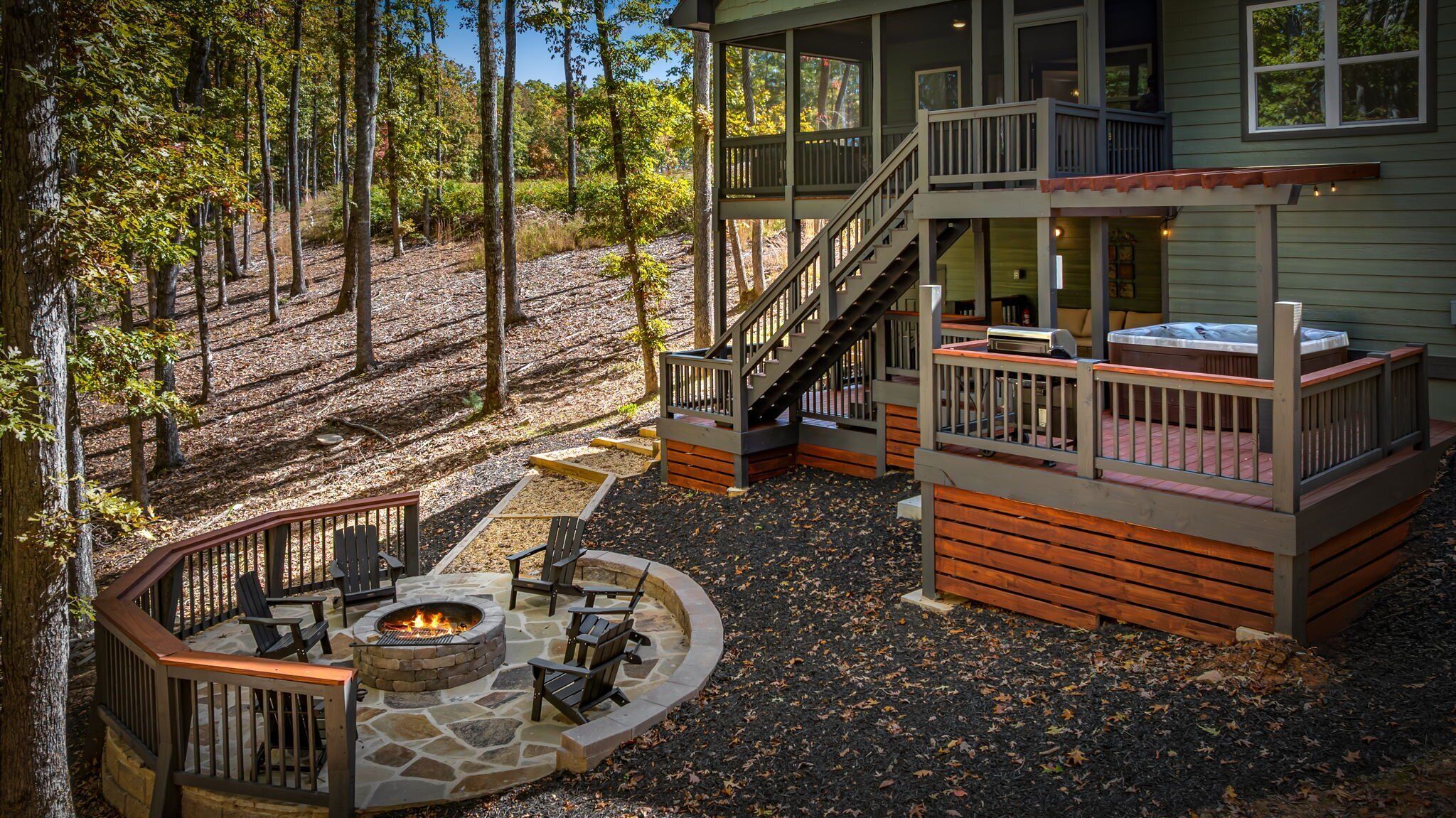 A two-story cabin with a stone fire pit and outdoor seating, surrounded by a forest.