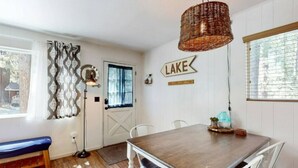 A dining area with a wooden table and white chairs, a wicker pendant light, and a sign that reads "LAKE". - Sunny Retreat | Near Beaches & Trails | Sleeps 6 (South Lake Tahoe)