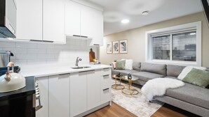 Una cocina moderna con armarios blancos, un lavamanos y una estufa. Hay un sofá gris con una manta blanca y una mesita de noche con una lámpara. Se ve una ventana con persianas en el fondo.