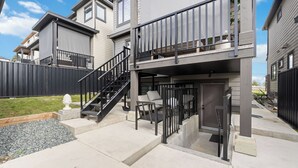 Un edificio residencial de dos pisos con un balcón, una escalera y un pequeño patio con mobiliario exterior.
