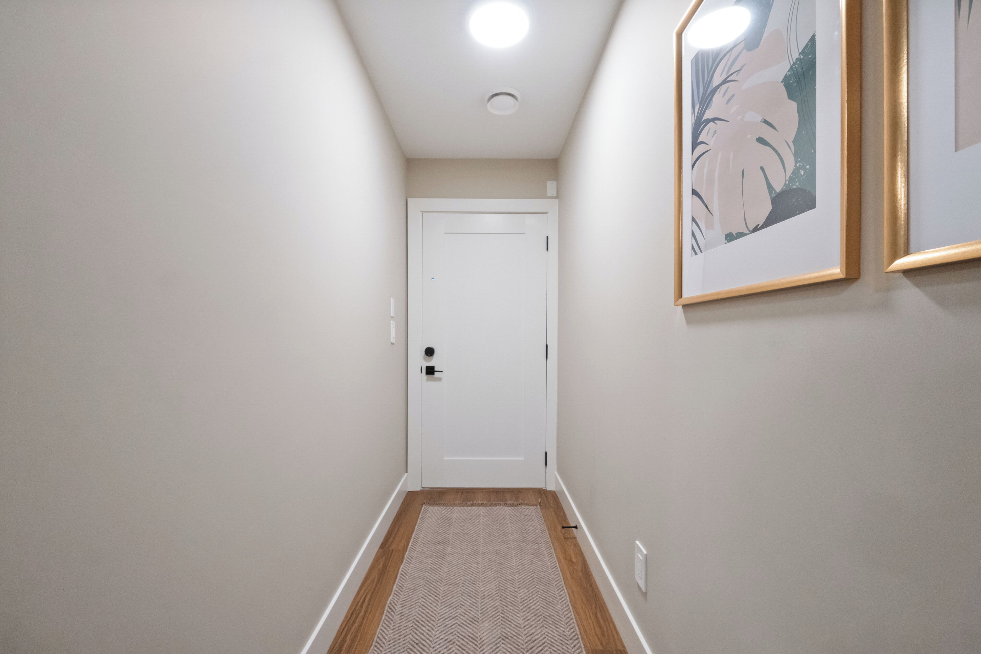Un couloir étroit doté d’un plancher en bois, une porte blanche au fond et deux tableaux encadrés accrochés au mur.