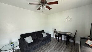 Un salon avec un canapé gris foncé, une table basse en verre, une table à manger avec des chaises, un ventilateur de plafond et une petite table d’appoint sur laquelle est posée une plante.