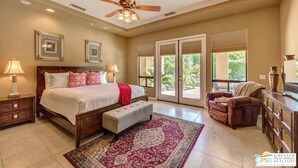 A bedroom with a bed, bedside tables, a ceiling fan, a large window, and a view of greenery outside. - Coachella Walkable | Tuscan Villa | Pool Sleeps 12 (Indio)
