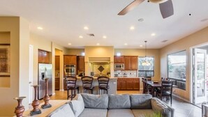 A modern living room with a gray sofa, wooden chairs, and a dining area with a table and chairs. A kitchen with wooden cabinets and appliances is visible in the background. - Coachella Walkable | Tuscan Villa | Pool Sleeps 12 (Indio)