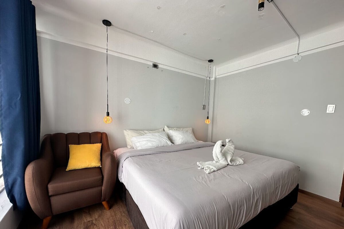 Una habitación de hotel moderna con una cama, un sillón marrón con un cojín amarillo y una mesita de noche.