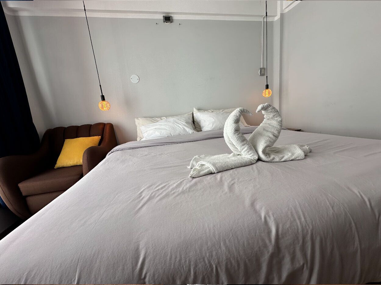 A neatly made bed with a towel sculpture, a brown armchair with a yellow pillow, and two hanging light bulbs.