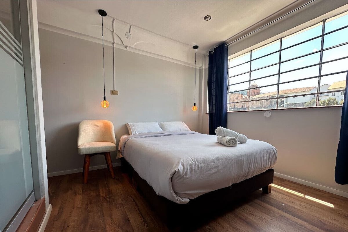 Un dormitorio moderno con una cama grande, una silla y vistas a un paisaje urbano a través de una ventana.