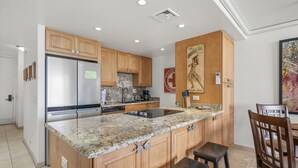 A modern kitchen with granite countertops, stainless steel appliances, wooden cabinets, and a bar area with stools. - Kahana Reef 210 (Lahaina)