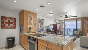 A modern kitchen with granite countertops, wooden cabinets, and stainless steel appliances. There is a dining area with a table and chairs, and a balcony with a view of the ocean. - Kahana Reef 210 (Lahaina)