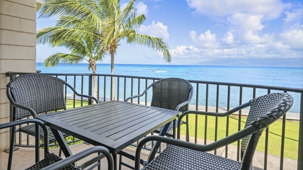 A balcony with a table and chairs, overlooking a beach with palm trees and the ocean. - Kahana Reef 210 (Lahaina)