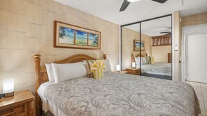 A bedroom with a large bed, wooden headboard, bedside tables with lamps, a ceiling fan, and a large mirror. - Kahana Reef 210 (Lahaina)