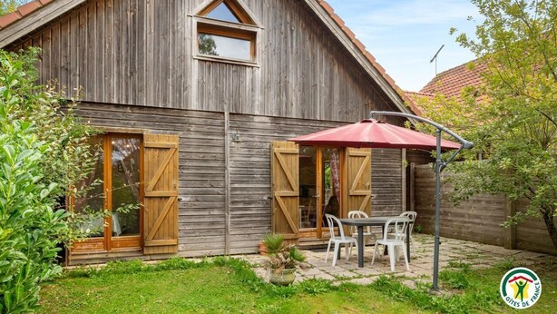 Un bâtiment rustique en bois, doté d’un toit en pente, de grandes fenêtres et d’un espace patio avec une table et des chaises.