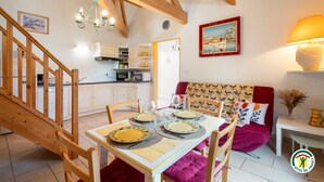 Un coin repas avec une table dressée pour quatre personnes, un escalier, une cuisine et un salon avec un canapé et un tableau.