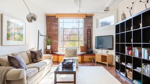 Una sala de estar con sofá, una mesa de centro, una estantería, televisor y un ventanal con vistas a árboles.