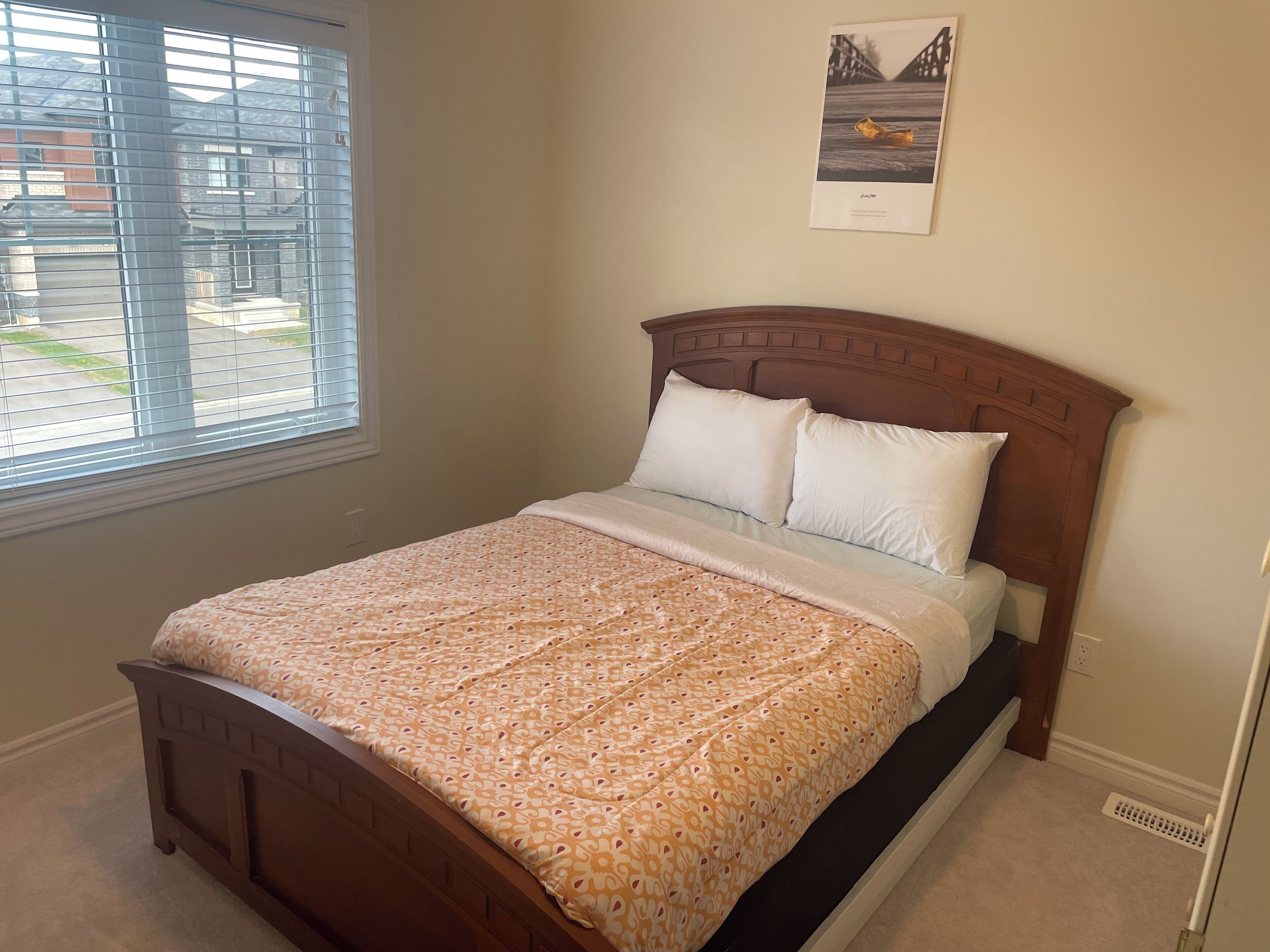 A bedroom with a wooden bed, a patterned bedspread, a window with blinds, and a framed picture on the wall.