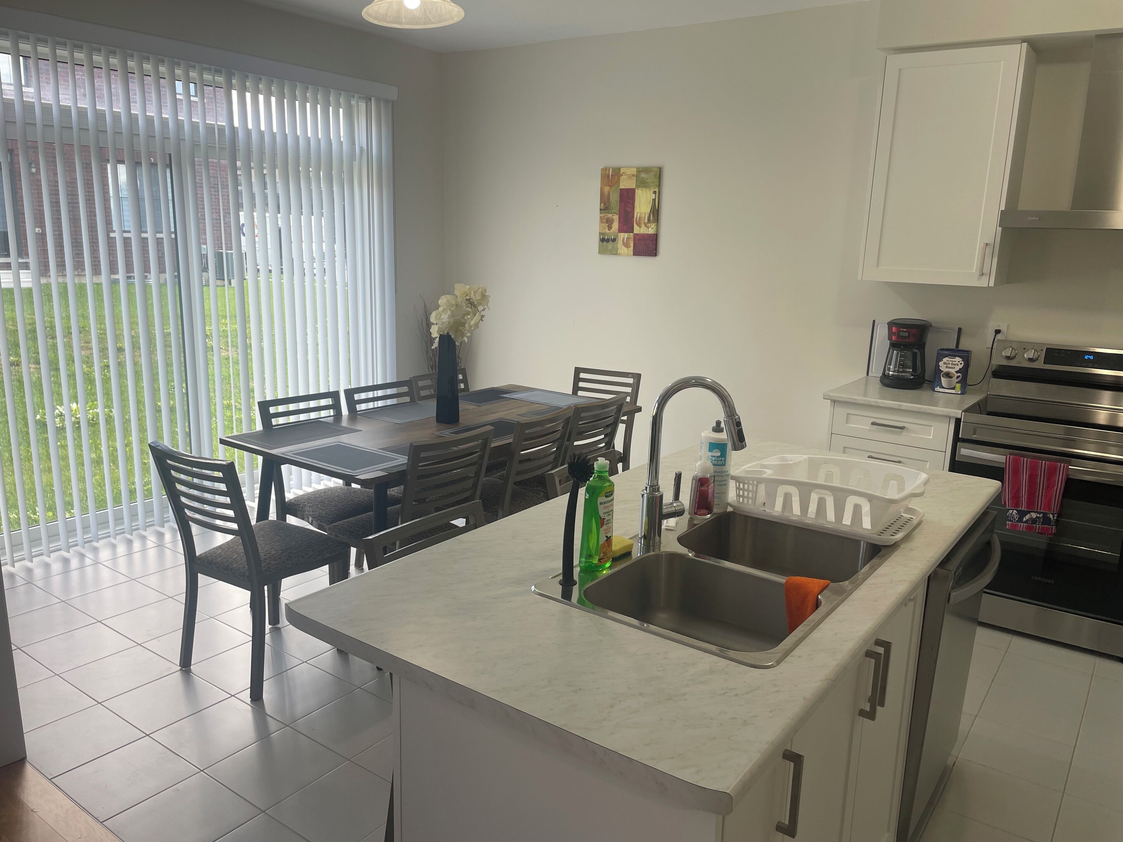 A modern kitchen with a dining area, a large window with blinds, and a stainless steel sink.