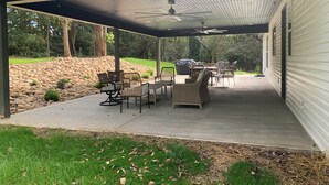 Een overdekt terras met buitenmeubilair, een plafondventilator en uitzicht op een stenen muur en bomen.
