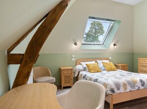 Ein Schlafzimmer mit geneigter Decke, einem Dachfenster, einem Bett mit gemusterten Textilien, einem Sessel, einem Nachttisch aus Holz und einem Kleiderschrank.
