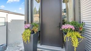Ein moderner Gebäudeeingang mit einer dunklen Tür, zwei großen Pflanzkübeln mit Blumen und blauem Himmel.
