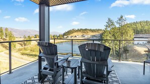 Ein Balkon mit zwei Adirondack-Stühlen und einem kleinen Tisch, mit Blick auf einen See und bewaldete Hügel.