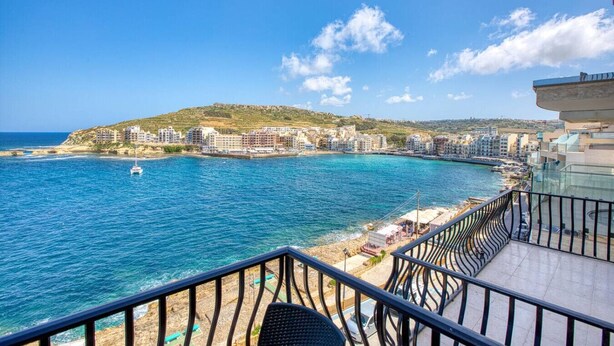 Un hotel sulla costa con un balcone vista mare, una spiaggia sabbiosa e uno sfondo collinare.