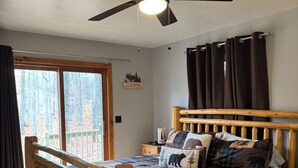 A wooden bed with a quilt, a ceiling fan, a window with curtains, and a nightstand with a lamp. - Spacious Cabin in LOTN- Great for ORV, Snowmobile, Golf, Ski or family getaways! (Mancelona)
