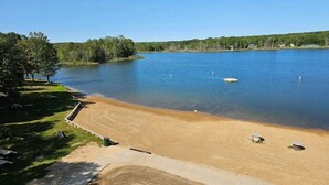 A sandy beach with a clear blue lake, surrounded by trees and a few buildings in the distance. - Spacious Cabin in LOTN- Great for ORV, Snowmobile, Golf, Ski or family getaways! (Mancelona)