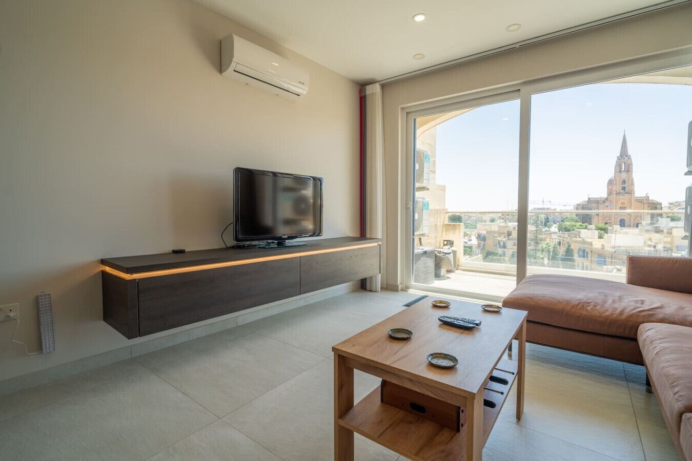 A modern living room with a flat-screen TV on a dark wooden console, a wooden coffee table, and a large window offering a city view.
