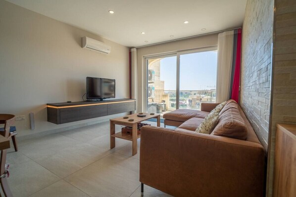 Un salon moderne avec un canapé, une table basse, un téléviseur posé sur une étagère intégrée et une grande fenêtre donnant sur des immeubles.
