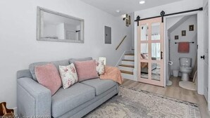 Una sala de estar con un sofá gris, un espejo, una escalera y un baño visible a través de una puerta abierta.
