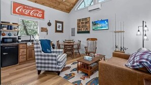 A cozy living room with a checkered sofa, wooden coffee table, and a dining area with a wooden table and chairs. A kitchen area with a refrigerator and microwave is visible, along with a Coca-Cola sign and motivational wall art.