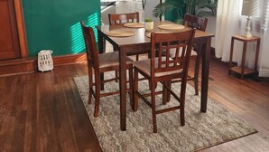 Uma área de refeições com uma mesa de madeira, cadeiras e um tapete. Cozinha ao fundo, com armários brancos e uma geladeira.